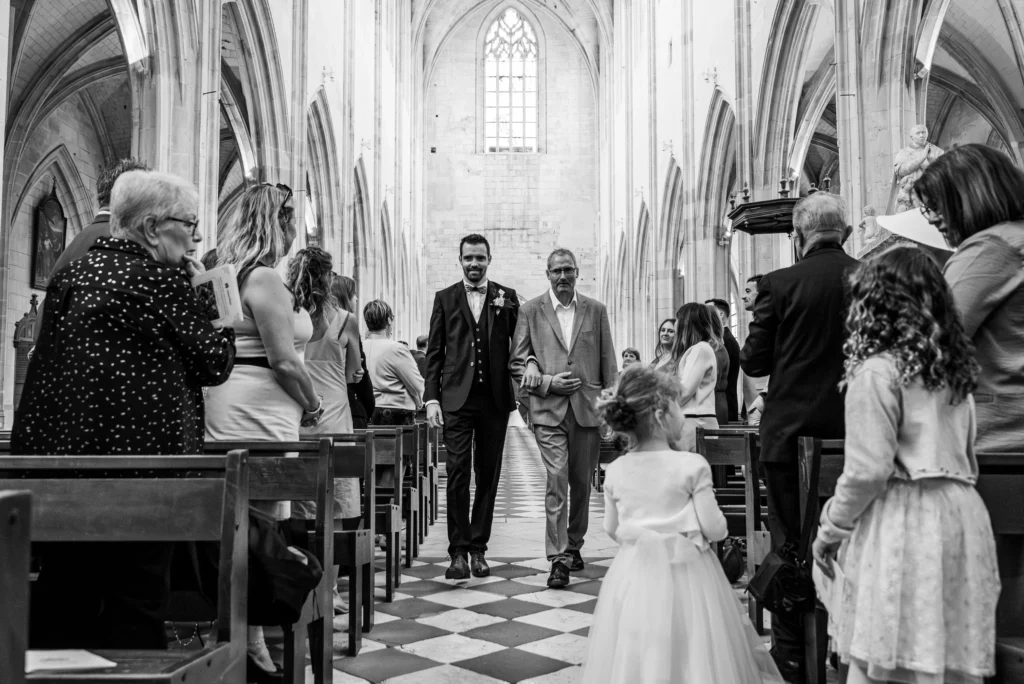 Photo du marié se dirigeant vers l'hotel au bras de son papa, entouré des invités lors de la cérémonie, photo prise par Quentin Viel